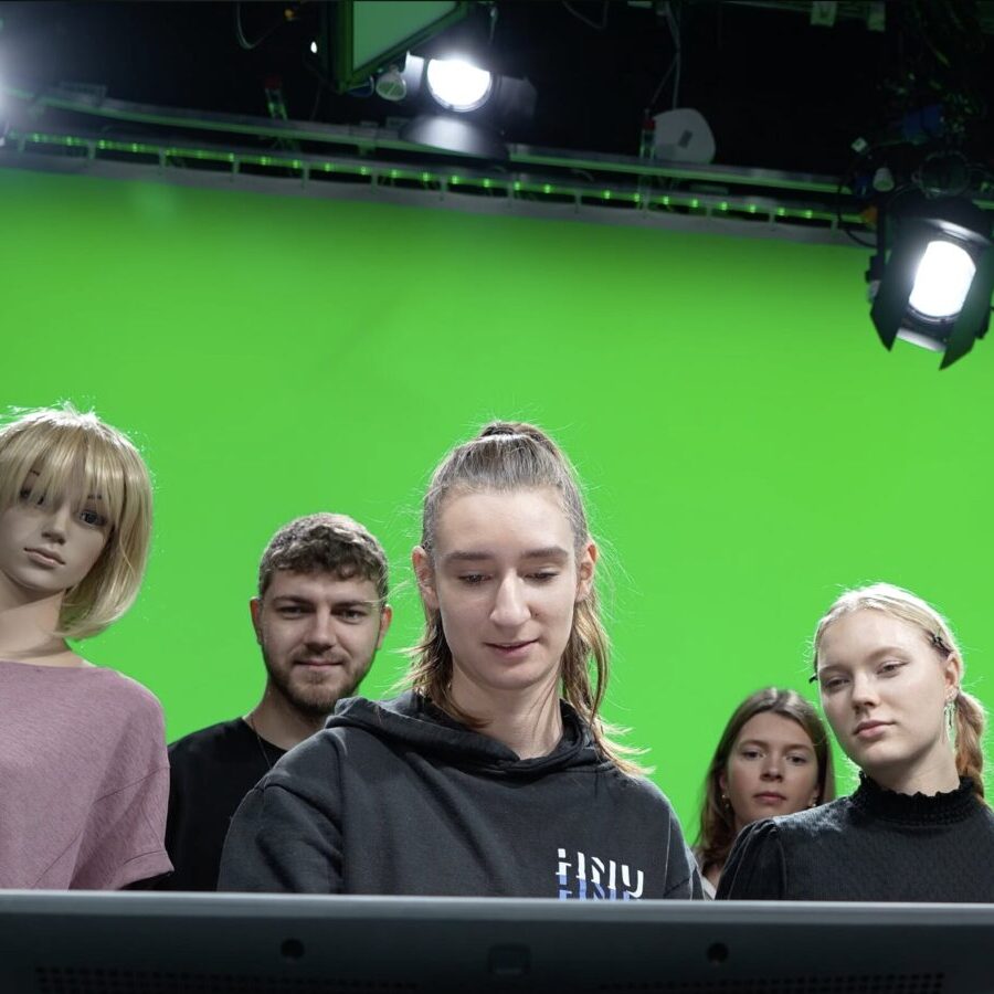 Gruppe von Studierenden steht vor einer großen Green-Screen-Wand und blickt auf einen Monitor; Studioleuchten hängen an der Decke, links steht eine Schaufensterpuppe.