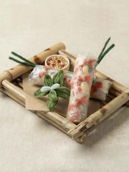 Stillleben: mehrere „Frühlingsrollen“ aus textilen Materialien auf einem Bambustablett, mit kleinen Dekoelementen.