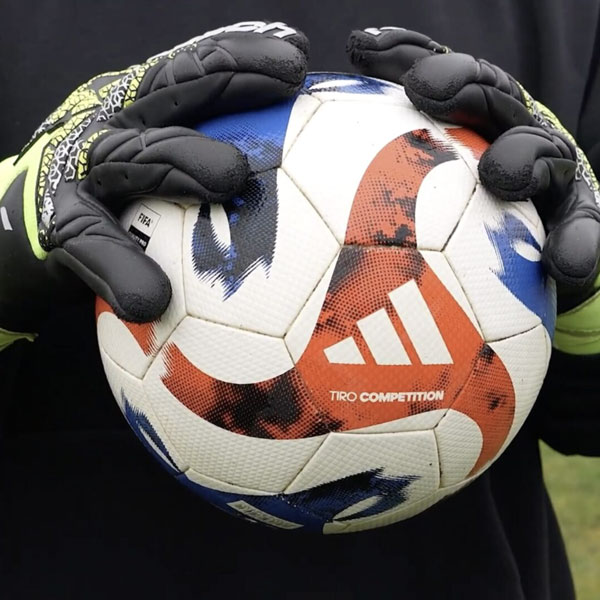 Nahaufnahme von Torwarthandschuhen, die einen Adidas-Fußball auf einem Fußballplatz festhalten.